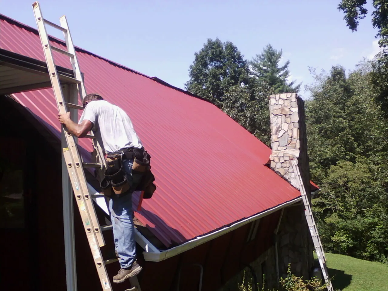Roofer on a ladder beside a red metal roof and stone chimney — Soffit & Fascia Repair by Roofing Brothers in Woodleaf